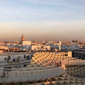 People walk along a modern, undulating walkway above a sprawling cityscape during a golden sunset.