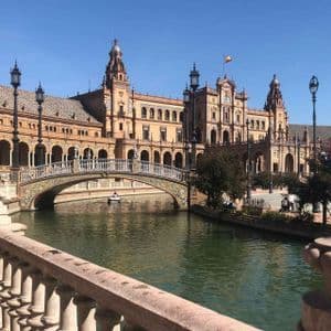 A view from behind a balustrade of an ornate building with a canal, where a person rows a boat under a tiled bridge.