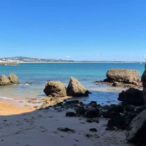 A view from a coastal cave looking out onto a sandy beach with large rocks in the turquoise sea under a clear blue sky.