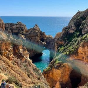 Un panorama da un sentiero a picco sul mare, con faraglioni dorati e un arco naturale su acqua turchese, e persone che passeggiano.