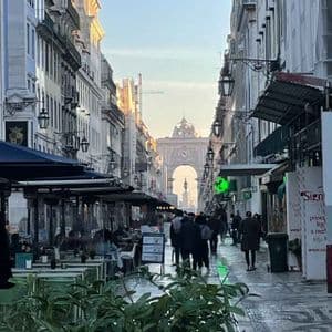 A pedestrian street lined with outdoor cafes and people walking towards a large triumphal arch in the distance.