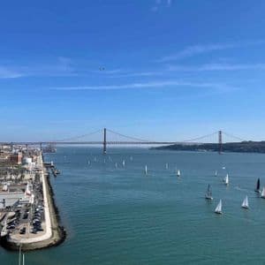 A panoramic view of sailboats on a wide river next to a city marina, with a large red suspension bridge in the background.