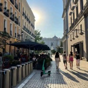 Quattro persone di un gruppo WeRoad camminano lungo una soleggiata strada acciottolata fiancheggiata da edifici, con un palazzo visibile in lontananza.