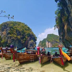 Eine Reihe traditioneller Langheckboote ankert an einem Sandstrand in einer tropischen Bucht mit großen, baumbestandenen Kalksteinfelsen.