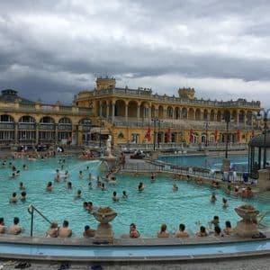 Molte persone si rilassano in una grande piscina termale all'aperto di fronte a un imponente edificio giallo sotto un cielo nuvoloso.