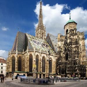 Una grande cattedrale gotica, con un tetto di tegole colorate e decorate e un'alta guglia, vista da una piazza acciottolata in una giornata parzialmente nuvolosa.