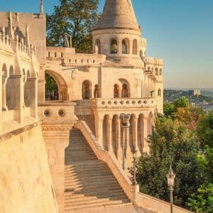Due persone sono sedute ai piedi di un'ampia scalinata in pietra che conduce a un bastione ornato con torri, immerso nella luce dorata del mattino.