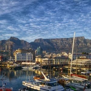 Un port rempli de bateaux amarrés avec une ville et une grande montagne à sommet plat en arrière-plan sous un ciel bleu texturé.