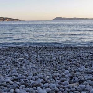 Ein Kiesstrand mit glatten grauen Steinen trifft auf das ruhige Meerwasser, wobei ferne Klippen am Horizont sichtbar sind.