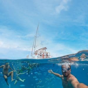 Eine WeRoad Gruppenreise schnorchelt unter Wasser zwischen Fischen, mit einem Mann, der ein Selfie macht, und einem Katamaran an der Oberfläche.