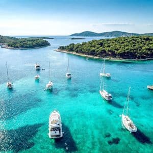Eine Luftaufnahme von Segelbooten und Yachten, die in einer ruhigen türkisfarbenen Bucht vor Anker liegen, umgeben von grünen, bewaldeten Inseln.