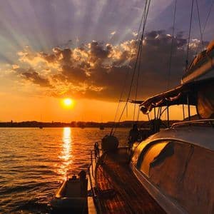 Blick von einem Segelboot auf einen goldenen Sonnenuntergang über dem Wasser, mit Sonnenstrahlen, die durch die Wolken brechen.