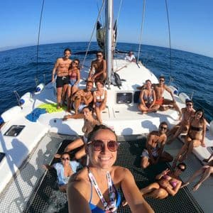 Eine WeRoad-Gruppe lächelt für ein Selfie auf dem Deck und im Netz eines Katamarans auf offener See unter einem klaren blauen Himmel.