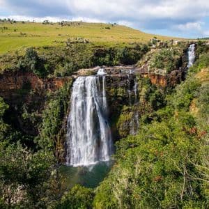 A large waterfall cascades over a rocky cliff into a pool of water, surrounded by dense green trees and rolling hills.