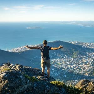Una persona en la cima de una montaña rocosa, con los brazos extendidos, contempla una ciudad costera y el océano.