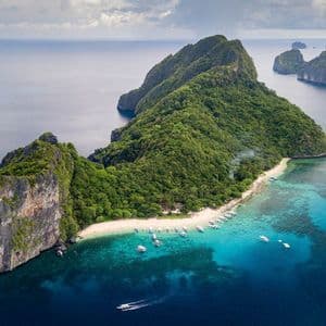 Una vista aerea di un'isola lussureggiante e rocciosa con una spiaggia di sabbia bianca dove molte piccole barche sono ormeggiate in acqua turchese.