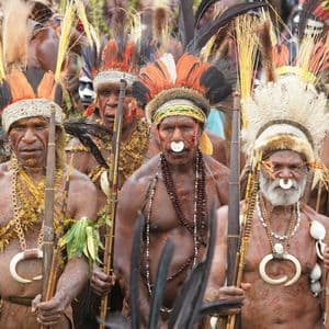 Un grupo de hombres indígenas con atuendos tradicionales, tocados de plumas, pintura corporal y joyas, portando lanzas.