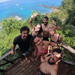 Un viaje en grupo de WeRoad se toma una selfie sonriente en un mirador de madera con vistas a una costa tropical de aguas azules.