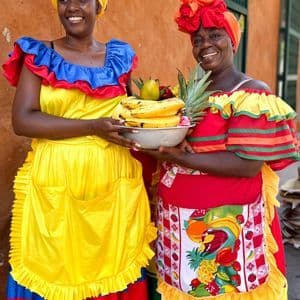 Zwei Frauen in farbenfrohen Trachten lächeln und balancieren Schalen mit Bananen und Ananas.