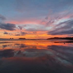 A colorful sunset with orange and pink clouds reflects on the wet sand of a beach, with small islands on the horizon.