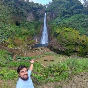 Un hombre barbudo se toma una selfie, apuntando a una imponente cascada que se precipita por un acantilado cubierto de musgo hacia el valle.