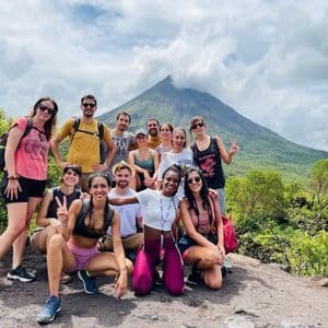 Un viaggio di gruppo WeRoad sorride per una foto su un sentiero sterrato con un grande vulcano coperto di nuvole e alberi verdi lussureggianti sullo sfondo.