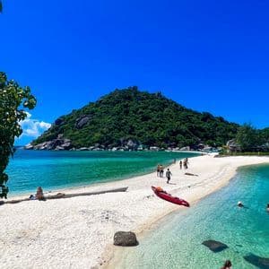 Des gens marchent sur un banc de sable blanc menant à une île verdoyante, avec un kayak rouge sur la plage et des nageurs dans l'eau turquoise cristalline.