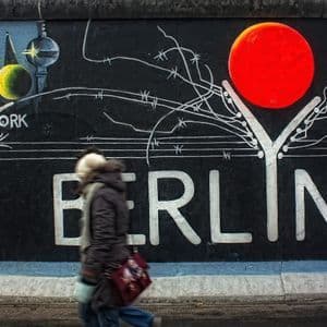 Una persona sfocata passa davanti a un grande murale di street art su un muro con la scritta 'BERLIN' in grandi lettere bianche.
