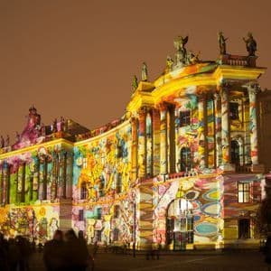 Una proiezione di luce colorata e astratta illumina di notte la facciata di un grande edificio classico, con spettatori in primo piano.