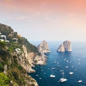 Una vista aerea di una costa rocciosa e verde con due faraglioni e numerose barche sull'acqua sotto un cielo rosa e arancione.