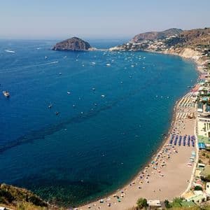 Vista aerea di una spiaggia sabbiosa affollata, con ombrelloni colorati lungo una baia di acqua blu intenso, barche e una cittadina costiera su una collina.