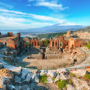 Un'ampia veduta delle rovine di un antico anfiteatro, con una città costiera e un vulcano innevato sullo sfondo.