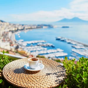 Una tazza di espresso su un tavolino di vimini che si affaccia su una baia costiera assolata con un porticciolo e una montagna distante.