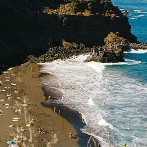 Una vista dall'alto di una spiaggia di sabbia nera dove le persone nuotano nell'oceano e prendono il sole accanto a una grande scogliera rocciosa.