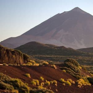 Un grande vulcano si erge su un paesaggio di terra vulcanica rossa e arbusti verdi sotto un cielo pallido e limpido.