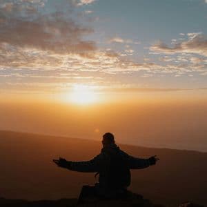 Una persona seduta su una roccia, con le braccia aperte, che osserva l'alba su un paesaggio di montagna.
