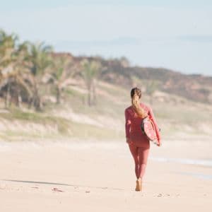 Una mujer en traje de neopreno rosa, vista de espaldas, camina por una playa de arena llevando una tabla de surf.