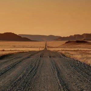 Une longue route de gravier traverse un paysage désertique vers des montagnes lointaines sous un ciel chaud et orangé.