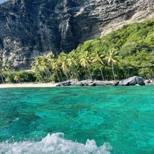 Una vista da una barca attraverso acque turchesi verso una spiaggia tropicale fiancheggiata da palme alla base di una grande scogliera rocciosa.