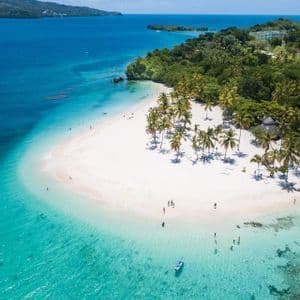 Una vista aerea di una spiaggia tropicale di sabbia bianca con palme, mentre le persone nuotano nell'oceano limpido e turchese.