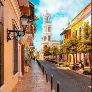 Una stretta strada acciottolata fiancheggiata da edifici colorati conduce a una torre dell'orologio bianca sotto un cielo blu con nuvole.
