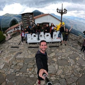 Un homme prend un selfie grand-angle avec son groupe WeRoad, posant autour d'une enseigne 'BOGOTA' sur une place en pierre.