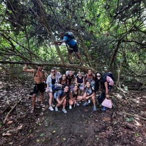 A WeRoad group trip with backpacks poses for a photo on a trail under tangled tree branches in a dense forest.