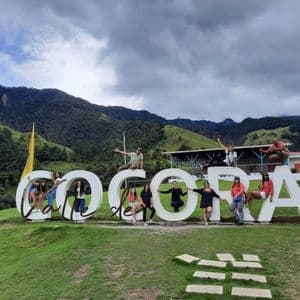 A WeRoad group trip posing for a photo on the large white 'Valle de Cocora' sign with a backdrop of green mountains.