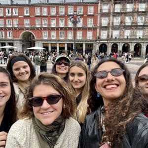 Un voyage de groupe WeRoad de femmes souriantes prenant un selfie sur une place animée d'une ville européenne avec des bâtiments historiques.