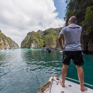 Un hombre con una camiseta de WeRoad de pie en la proa de un barco, contemplando una laguna turquesa rodeada de acantilados rocosos.