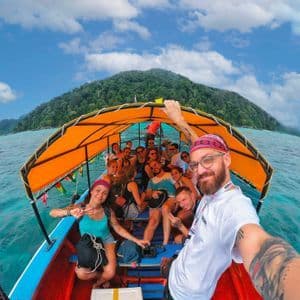 Un homme prend un selfie d'un voyage de groupe WeRoad sur un bateau avec un auvent orange, naviguant sur des eaux turquoise près d'une île luxuriante.