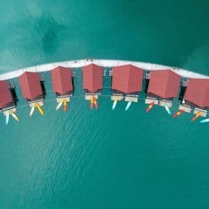 Luftaufnahme geschwungener schwimmender Bungalows mit roten Dächern und vertäuten Kajaks auf türkisfarbenem Wasser.