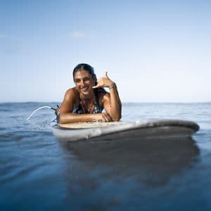 A woman lies on her surfboard in the ocean, smiling and making a shaka hand sign toward the camera.