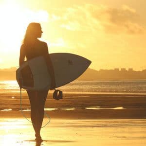 A person in silhouette holds a surfboard while standing on a wet beach at sunset, with the ocean and a distant shoreline in view.